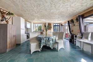 A spacious kitchen with a dining area, featuring a table with a patterned tablecloth, white chairs, and a view of the outdoors through large windows. - Charming Winegrower's House with Lake View, Private Pool, and Wi-Fi (Guizengeard)
