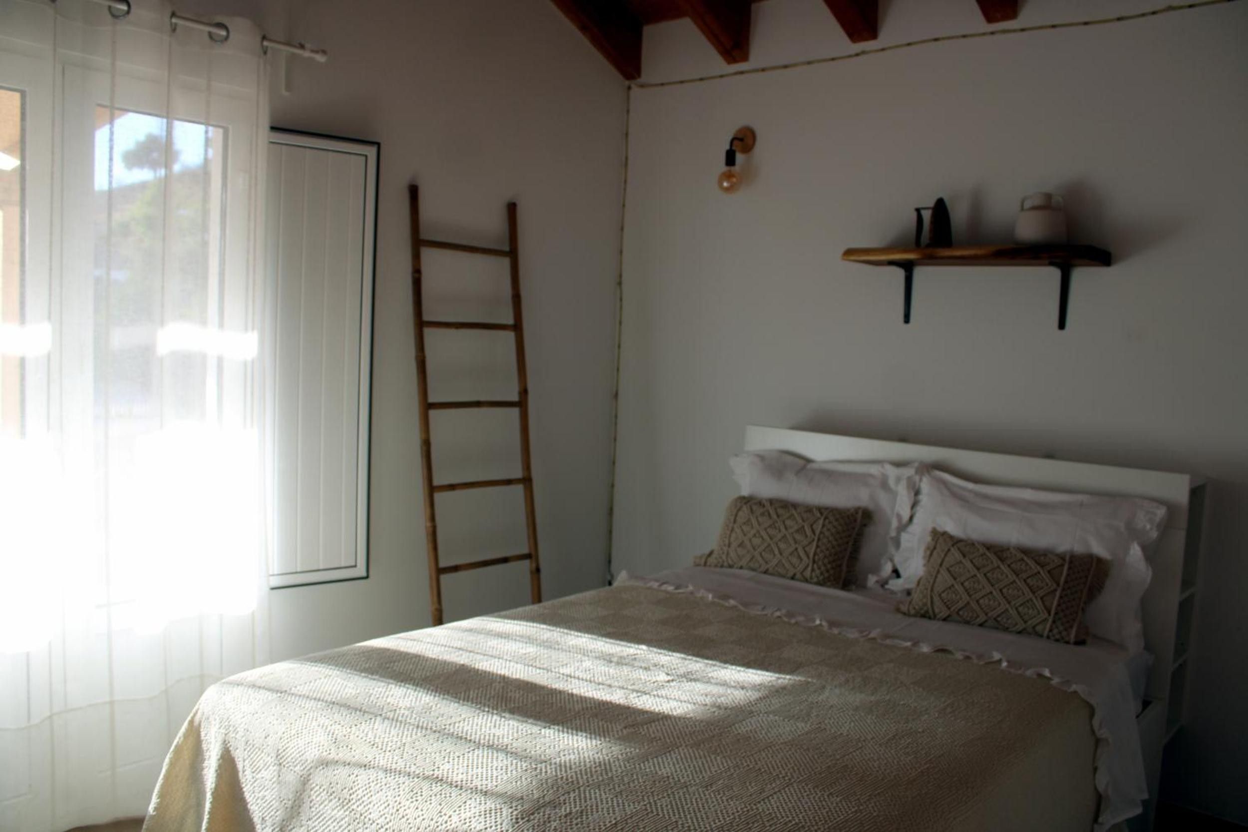 Ein Schlafzimmer mit einem Bett, einer Leiter, einem fensterbelaubten Fenster und einem an der Wand befestigten Regal mit Gegenständen.