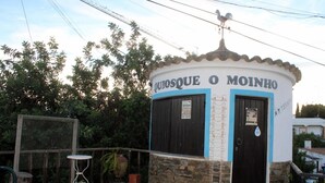 Um pequeno edifício redondo com uma placa que diz 'QUOSQUE O MOINHO'. Tem um telhado telhado, uma pequena porta e uma janela.