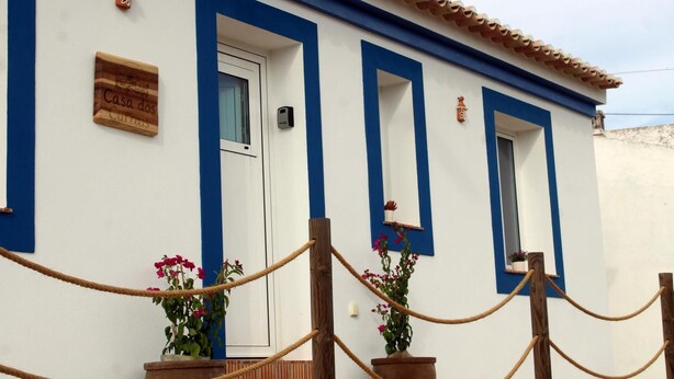 Um edifício com janelas e portas emolduradas em azul, uma placa com a inscrição 'Casa dos Currais' e vasos com plantas de flores cor-de-rosa.