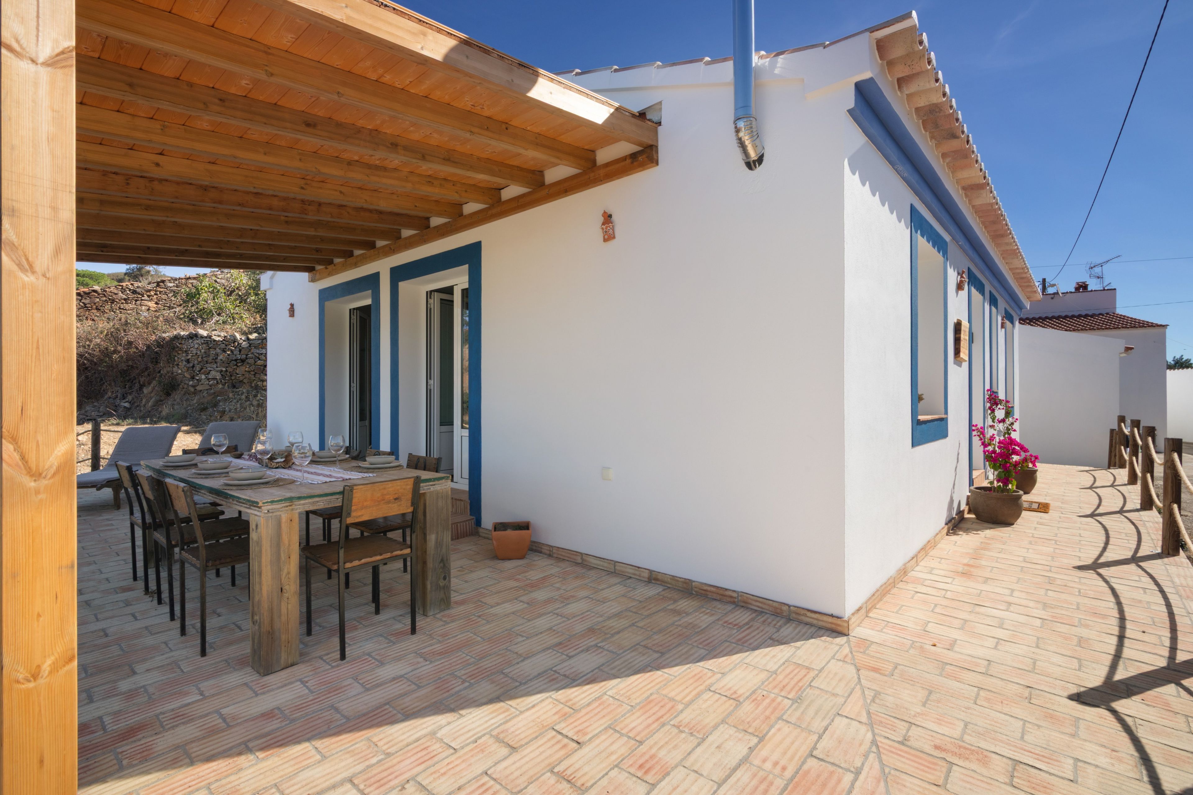 A covered patio with a wooden table and chairs, a stone wall in the background, and a white building with blue trim.