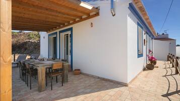 A covered patio with a wooden table and chairs, a stone wall in the background, and a white building with blue trim.