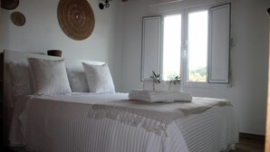 Cama bem arrumada com lençóis e travesseiros brancos, uma planta na mesa de cabeceira e uma janela vista.