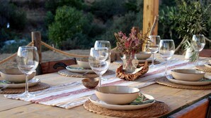 Mesa de jantar de madeira posta com pratos, tigelas e copos de vinho brancos, com um fundo rústico de folhagem e estrutura de madeira.