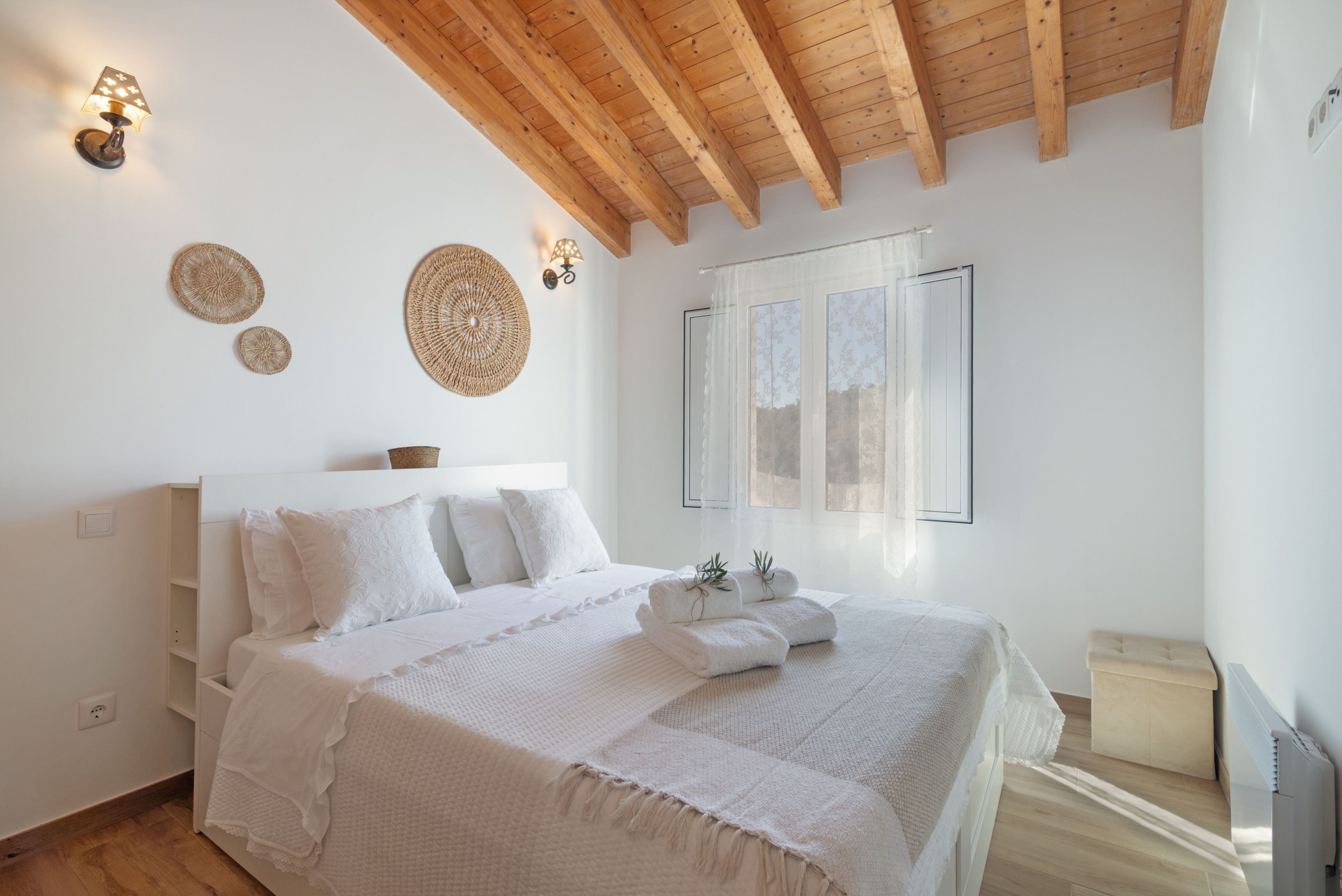 A bedroom with a wooden ceiling, a bed with white bedding, wall-mounted lamps, and a window with sheer curtains.