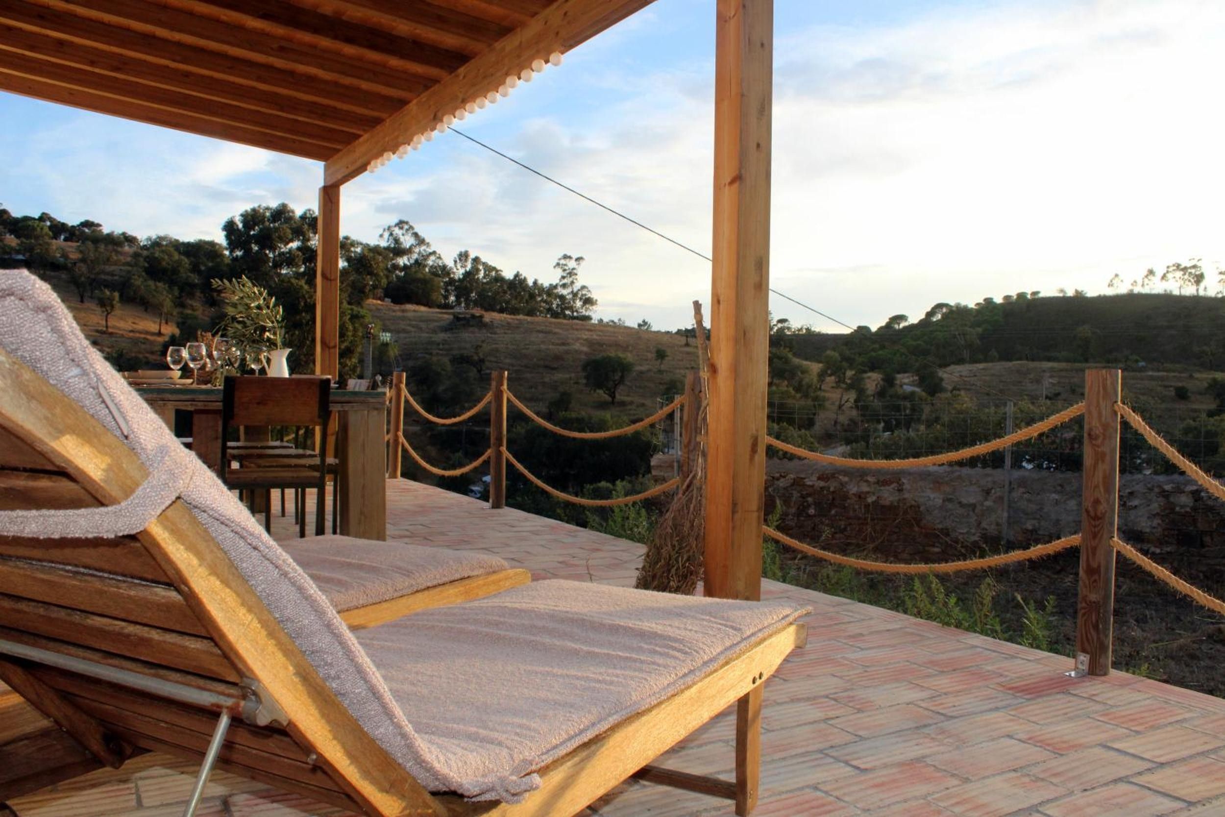 A wooden deck with a lounge chair, a table with a vase and glasses, and a view of a hilly landscape.