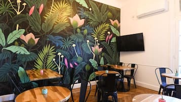 Interior de restaurante con mesas y sillas redondas, un mural grande de follaje tropical y un televisor de pantalla plana en la pared.