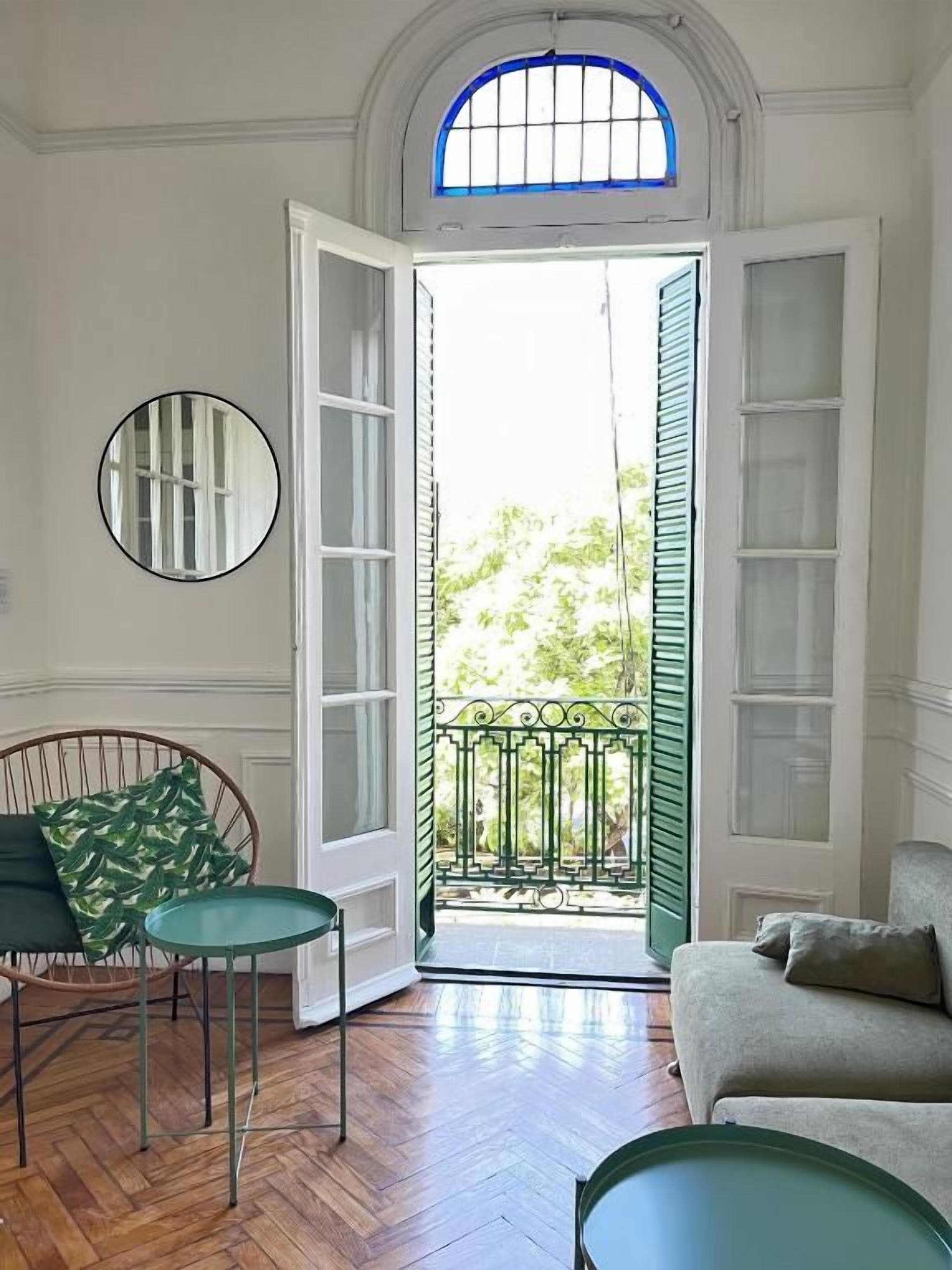 Una habitación con suelo de madera, una puerta verde, un espejo redondo, una silla verde y un sofá.