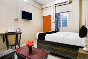 Habitación de hotel con una cama, un escritorio con silla, un televisor, un equipo de aire acondicionado y una ventana con cortinas.