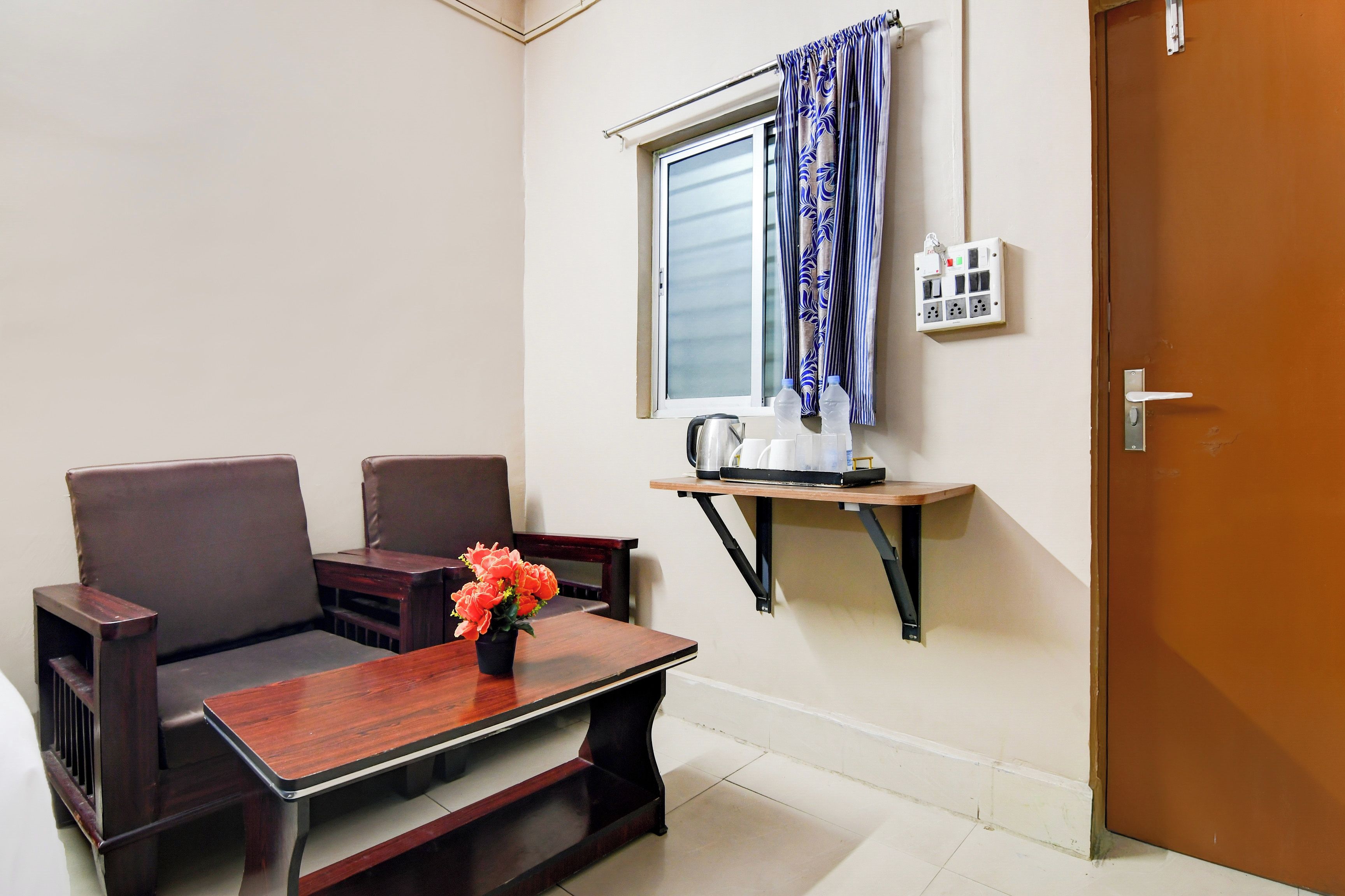 A small room with a sofa, a table with a vase of flowers, a window with curtains, a kettle, and a power outlet.
