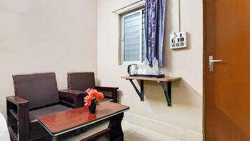 Una habitación pequeña con sofá, una mesa con un jarrón de flores, ventana con cortinas, hervidor eléctrico y toma de corriente.
