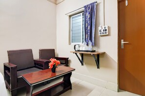 Una habitación pequeña con sofá, una mesa con un jarrón de flores, ventana con cortinas, hervidor eléctrico y toma de corriente.