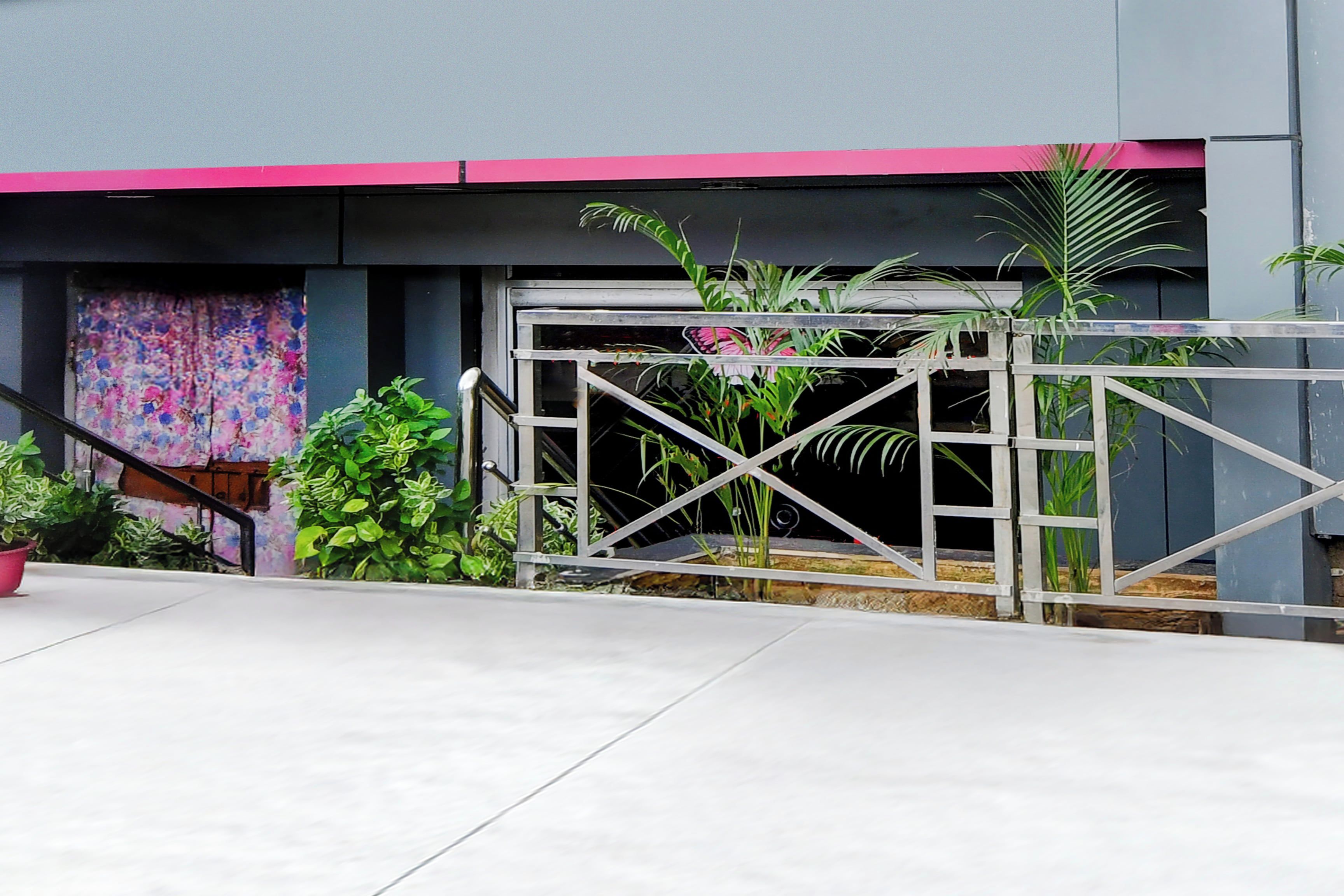 Entrada com portão de metal, plantas em vasos e escadaria que leva a um edifício com toldo rosa.