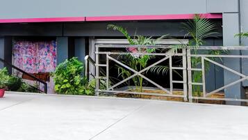Entrada com portão de metal, plantas em vasos e escadaria que leva a um edifício com toldo rosa.