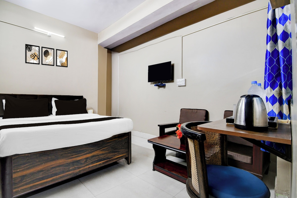A hotel room with a bed, a desk with a kettle, a chair, a TV, and framed artwork on the wall.