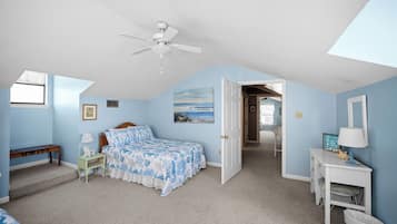 A bedroom with a bed, a desk, a chair, a ceiling fan, and a door leading to another room.