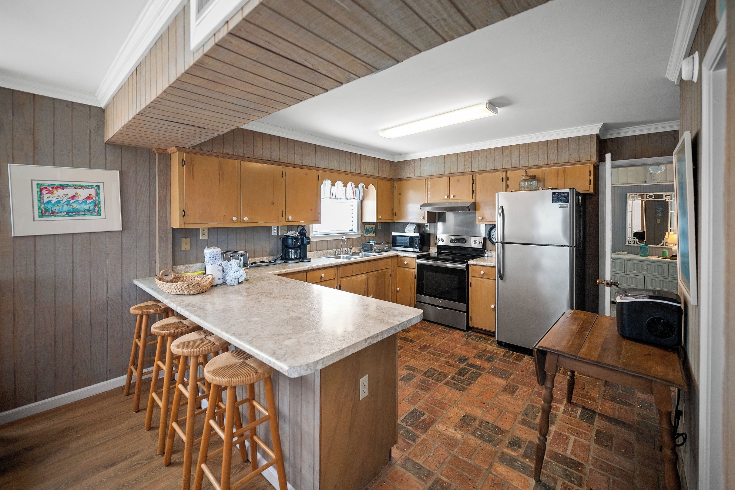 Una cocina moderna con armarios de madera, una isla central con taburetes de bar, un refrigerador de acero inoxidable y un suelo con estampado de ladrillo.