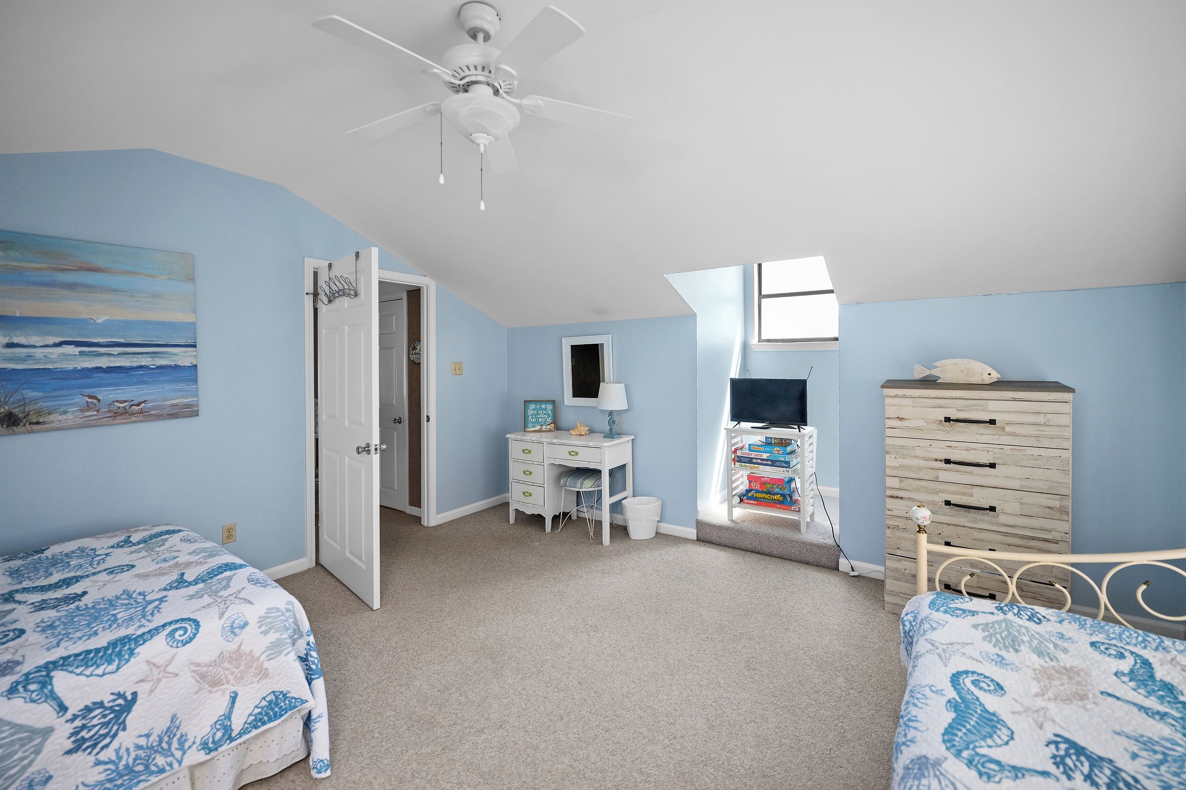 A bedroom with two beds, a desk, a dresser, a ceiling fan, and a beach-themed painting.