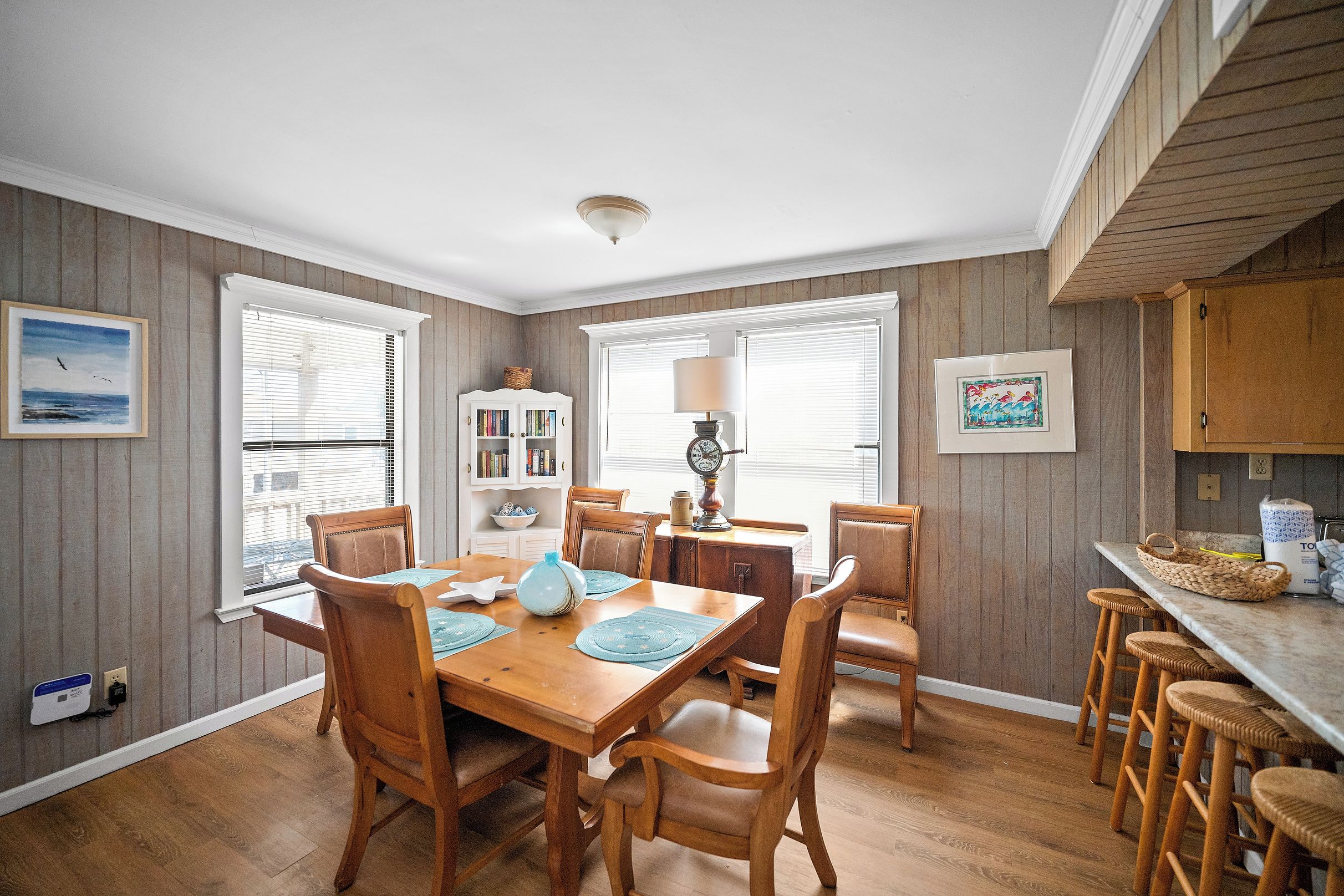 A dining area with a wooden table and chairs, a bookshelf, and a kitchen area with a counter and stools.