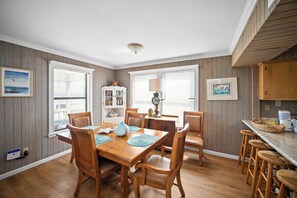 Een eethoek met een houten tafel en stoelen, een boekenkast en een keuken met een aanrecht en krukken.
