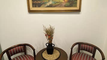 Een eethoek met een ronde tafel, twee gestoffeerde stoelen, een vaas met gedroogde bloemen en een lijst met een landschapsschilderij aan de muur.