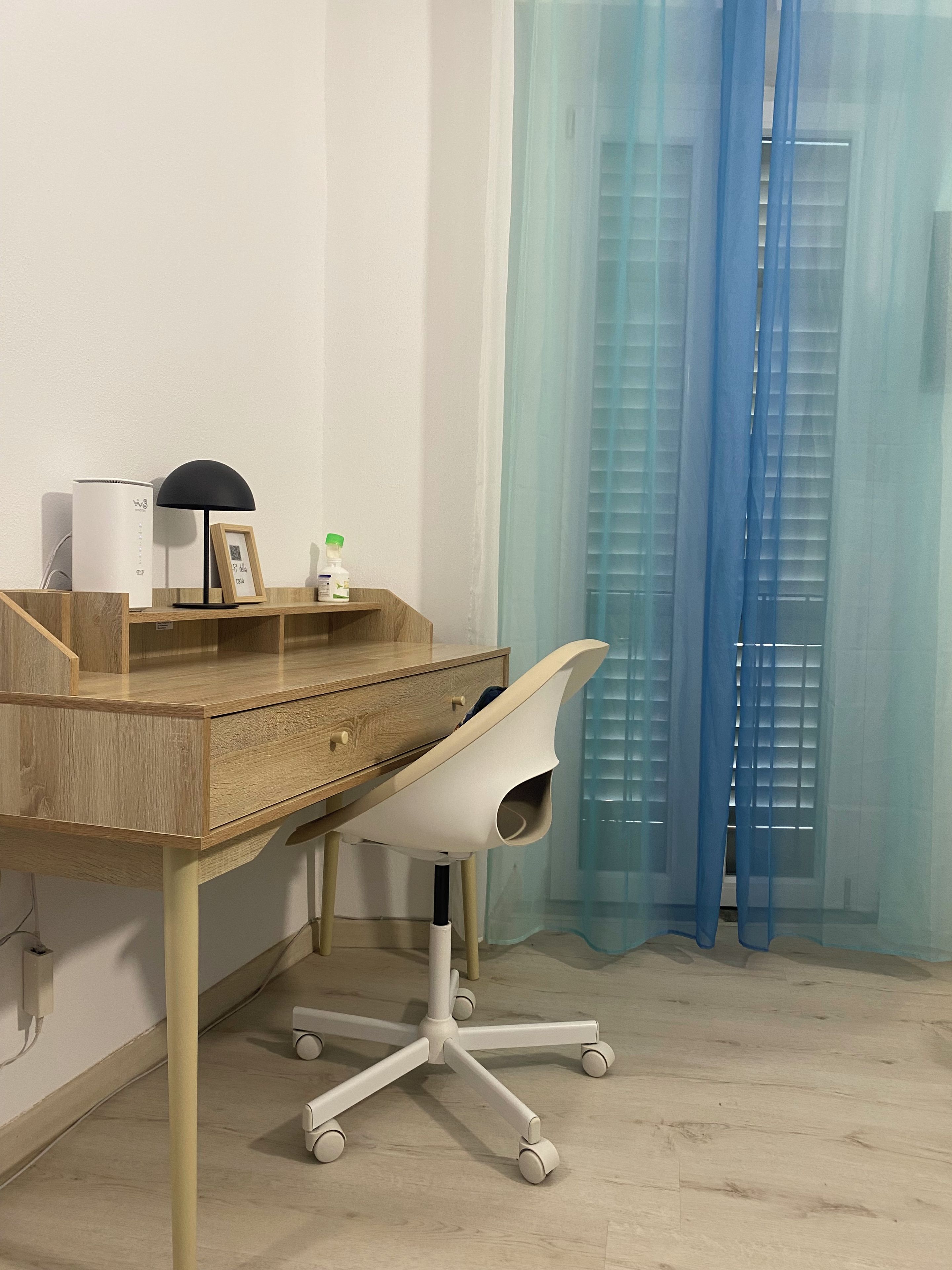 Een kamer met een houten bureau, een witte bureaustoel, een witte luchtreiniger, een zwarte bureaulamp, een lijst met een foto en een blauw gordijn.
