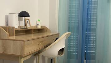 Een kamer met een houten bureau, een witte bureaustoel, een witte luchtreiniger, een zwarte bureaulamp, een lijst met een foto en een blauw gordijn.