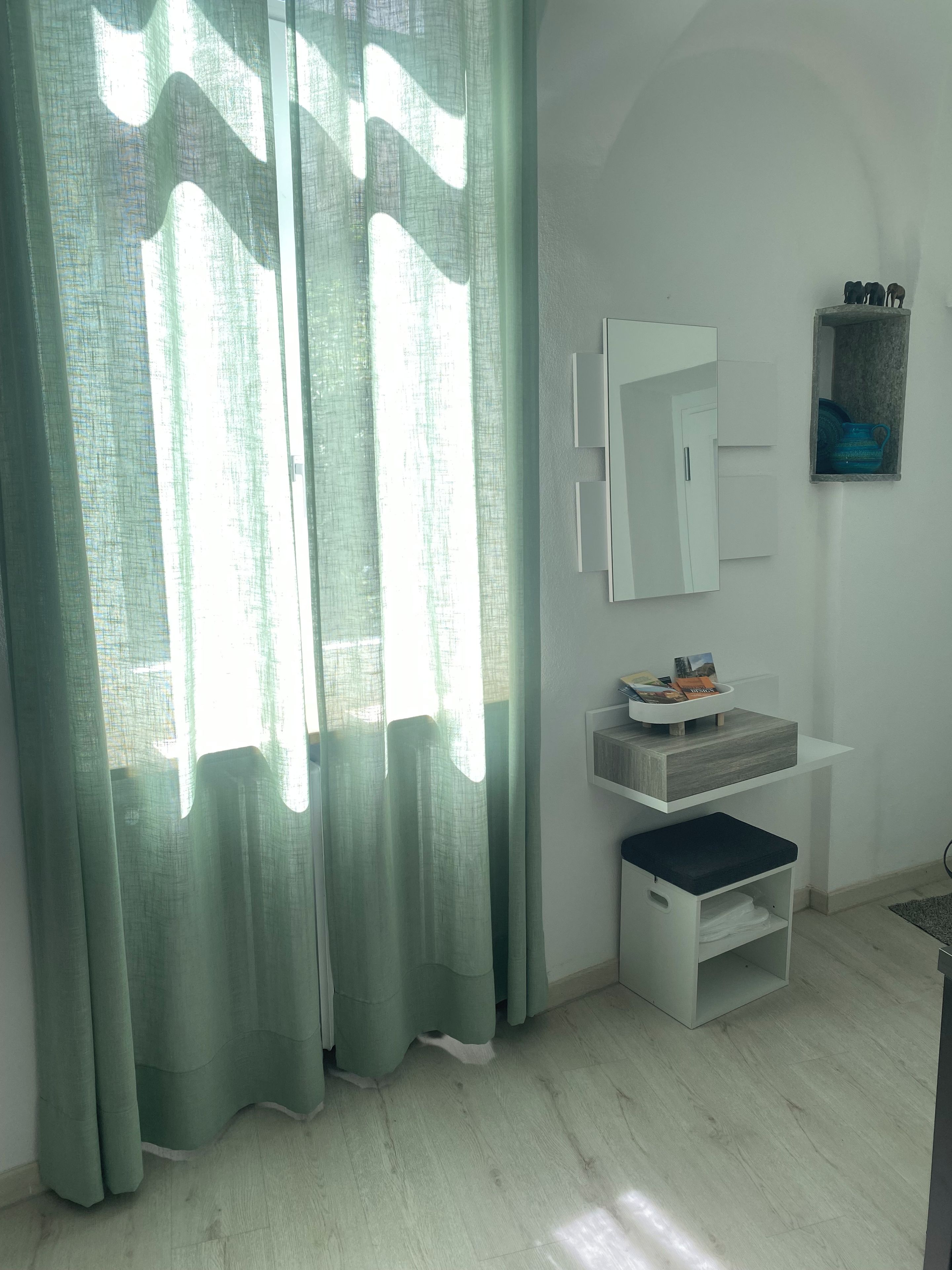 Ein Zimmer mit Fenster, Spiegel, Ablagefläche mit Gegenständen und einem kleinen Hocker.