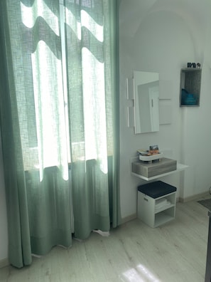 Une pièce avec une fenêtre, un miroir, une étagère avec quelques objets et un petit tabouret.