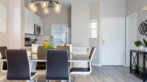Een moderne eethoek met een witte tafel en zwarte stoelen, een keuken op de achtergrond en een deur aan de rechterkant.
