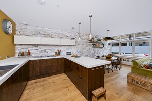 Een moderne keuken met een wit aanrecht, houten keukenkastjes en een bakstenen muur. Er is een zithoek met een groene bank en een rieten mand.