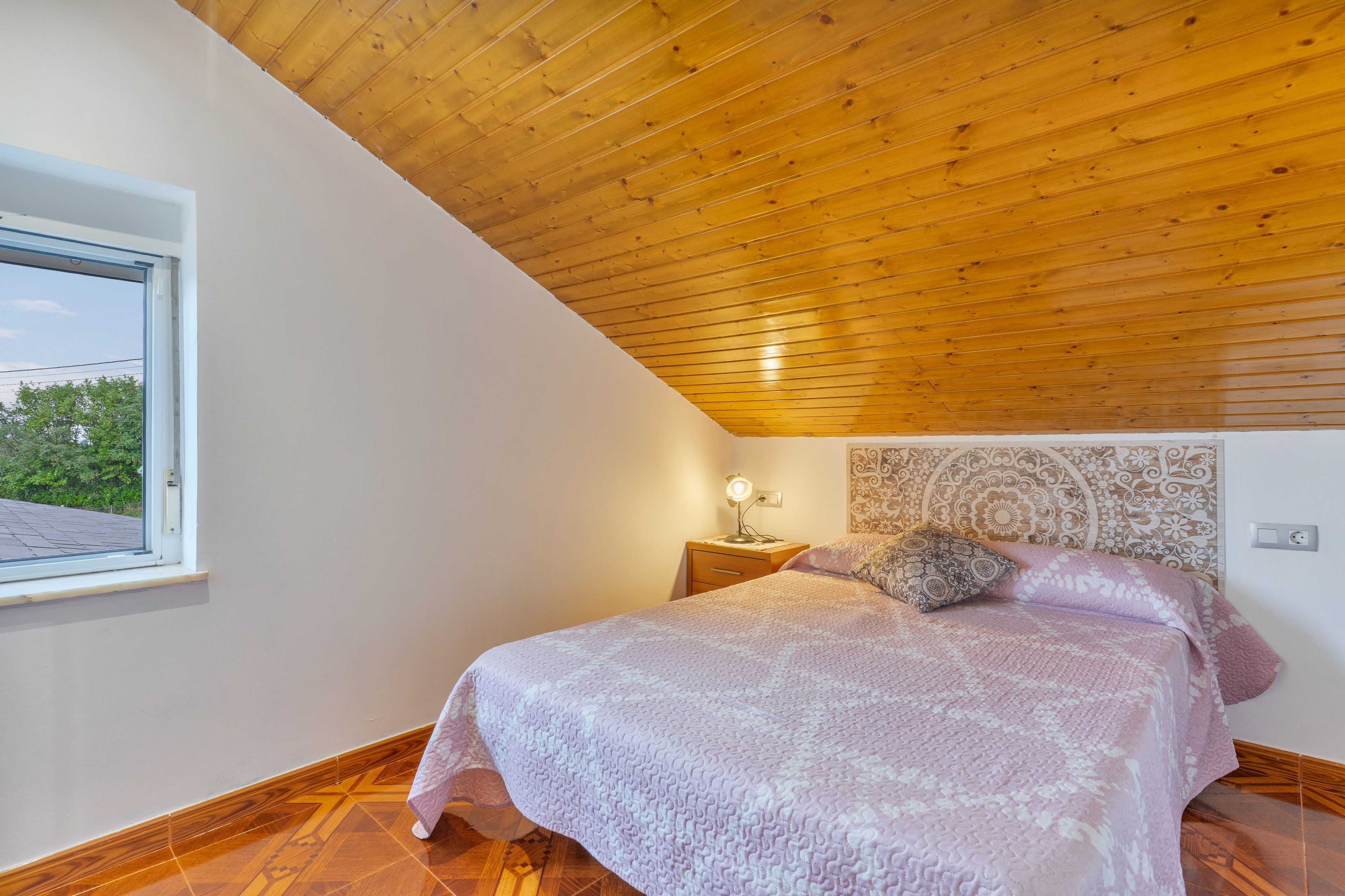 Een slaapkamer met een houten plafond, een bed met beddengoed met een patroon, een nachtkastje met een lamp en een raam met uitzicht op bomen.