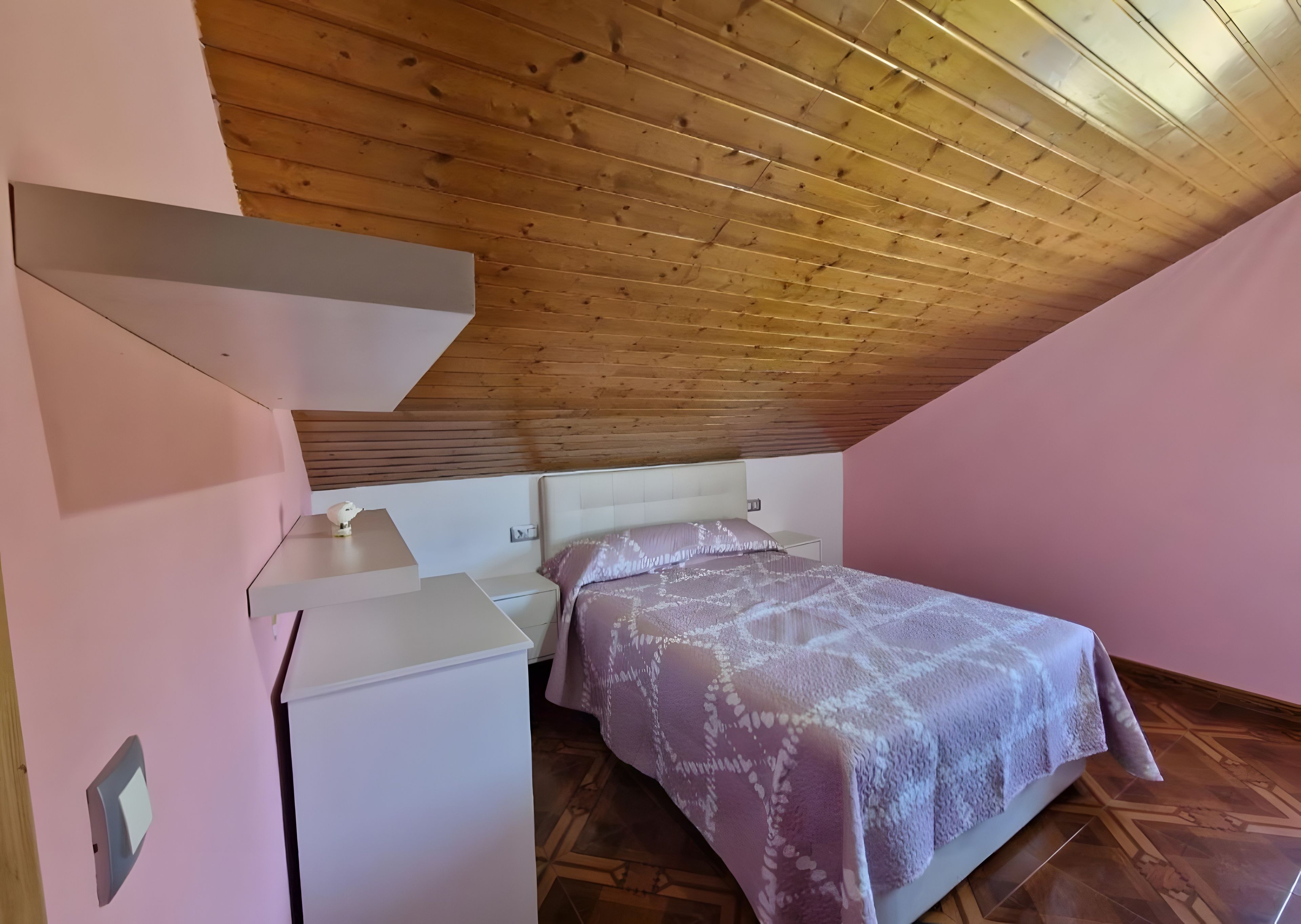Una camera da letto con soffitto a falde, un letto con coperta fantasia, un comodino e un tavolino con una lampada.