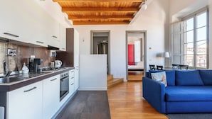 A modern kitchen with white cabinets and a wooden ceiling, a dining area with a blue sofa and a lamp, and a view of a red room through an open door. - Charming Apt in the Heart of Florence (Firenze)