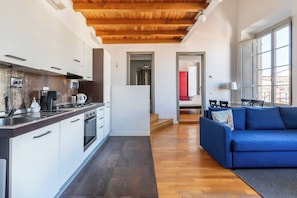 A modern kitchen with white cabinets and a wooden ceiling, a dining area with a blue sofa and a lamp, and a view of a red room through an open door. - Charming Apt in the Heart of Florence (Firenze)