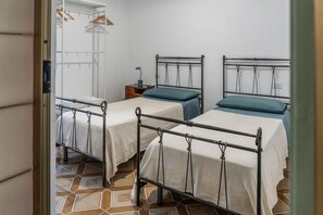 Ein Zimmer mit zwei Betten mit Metallgestell, einem kleinen Nachttisch und einem an der Wand befestigten Regal.