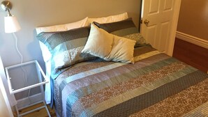 Una cama bien hecha con una colcha a rayas y dos almohadas, una lámpara de noche y una puerta al fondo.