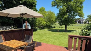 Een houten terras met een tafel en stoelen, een parasol en uitzicht op een verzorgd gazon en bomen.