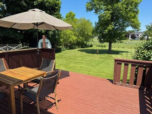 Une terrasse en bois avec une table et des chaises, un parasol, et une vue sur une pelouse bien entretenue et des arbres.
