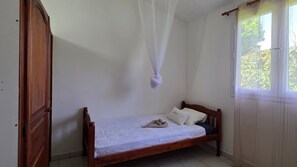 A single bed with a wooden frame and white bedding, a window with sheer curtains, and a closed wooden door. - Maison Bois Jadé - Proche de La Trinité (Arrondissement of La Trinité)