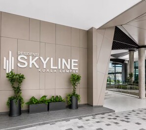 Une entrée moderne d’un immeuble, avec une enseigne indiquant « RESIDENSI SKYLINE KUALA LUMPUR » et des plantes en pot.