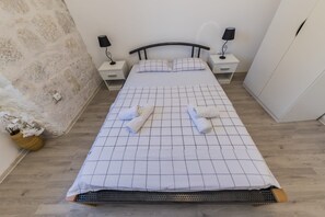 Een netjes opgemaakt bed met een bedspreider met ruitmotief, twee nachtkastjes met lampen, een houten nachtkastje en een vaas met witte bloemen.