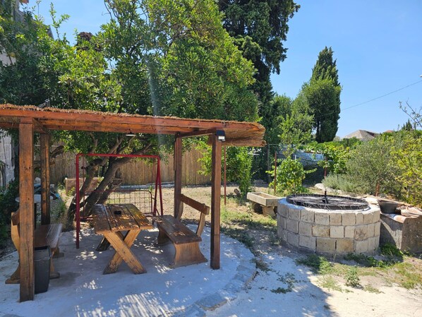 Ett rustikt utomhusområde med en träpergola, picknickbord, en eldstad av sten och en gunga.