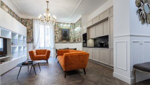 Ein modernes Wohnzimmer mit einem Kronleuchter, zwei orangefarbenen Sesseln, einem Couchtisch und einem Fernseher auf einer weißen Kommode.