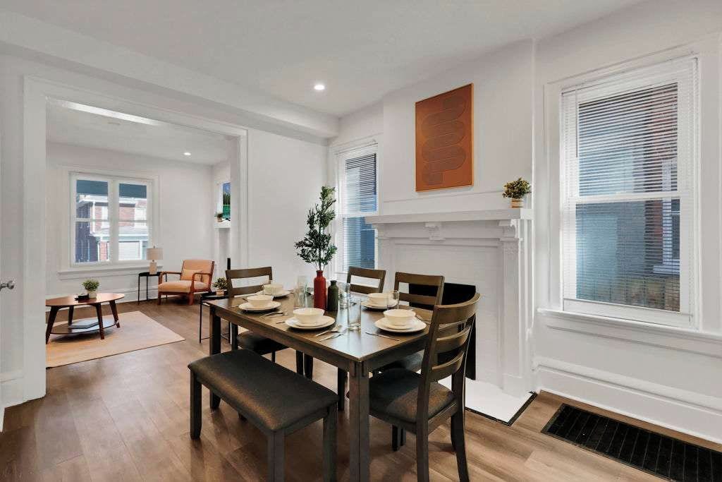 A dining area with a wooden table set for six, surrounded by chairs, with a fireplace and a large window with blinds.