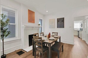 Uma sala de jantar com mesa de madeira, cadeiras e lareira. Há uma planta em vaso à esquerda e obras de arte nas paredes.