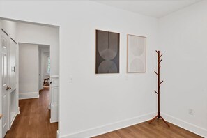 A hallway with white walls and wooden flooring, leading to a room with two framed artworks and a coat rack. - Olde Towne East 4BR Townhome Near East Market (Columbus)