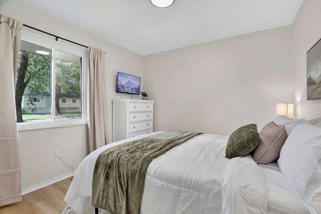 Un dormitorio con cama, televisor sobre un mueble, ventana con cortinas y un cuadro en la pared.