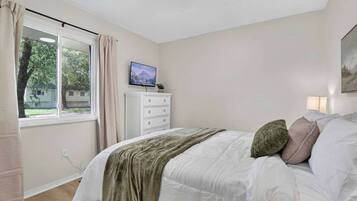 Un dormitorio con cama, televisor sobre un mueble, ventana con cortinas y un cuadro en la pared.
