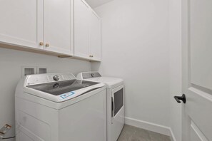 A laundry room with a front-loading washer and dryer, white cabinets above, and a door to the right. - FUNtastic Home! | 5 BR - Hot Tub + Game Room! (Columbus)
